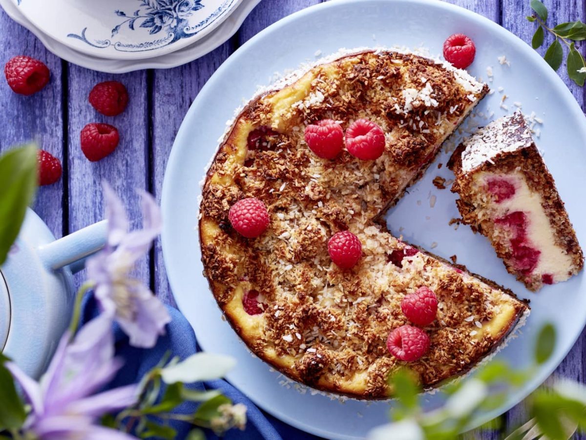 Ein Schmandkuchen mit Himbeeren auf einem hellblauen Teller auf einem hellblauen Holztisch. Auf dem Tisch sind Himbeeren, Geschirr und Besteck zu sehen.