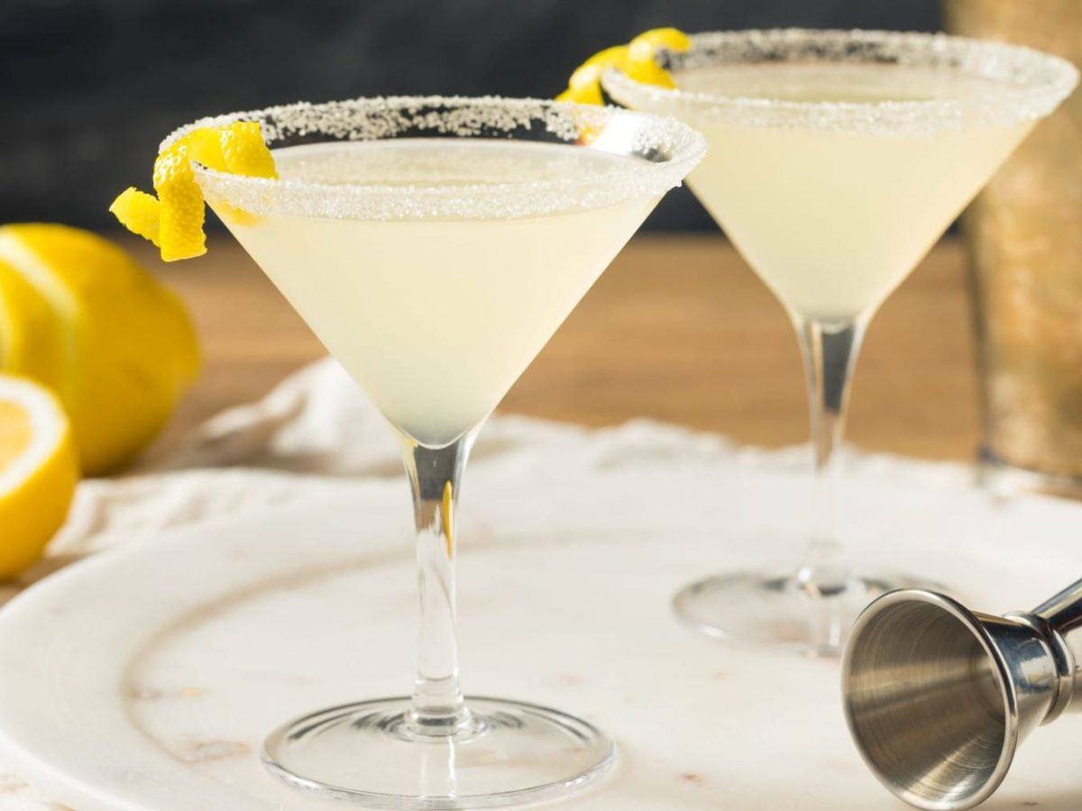 Zwei Lemon Drop Martini mit Barmaß auf hellem Teller, Tuch und Holztisch. Im Hintergrund Shaker und Zitronen. Frontalaufnahme.