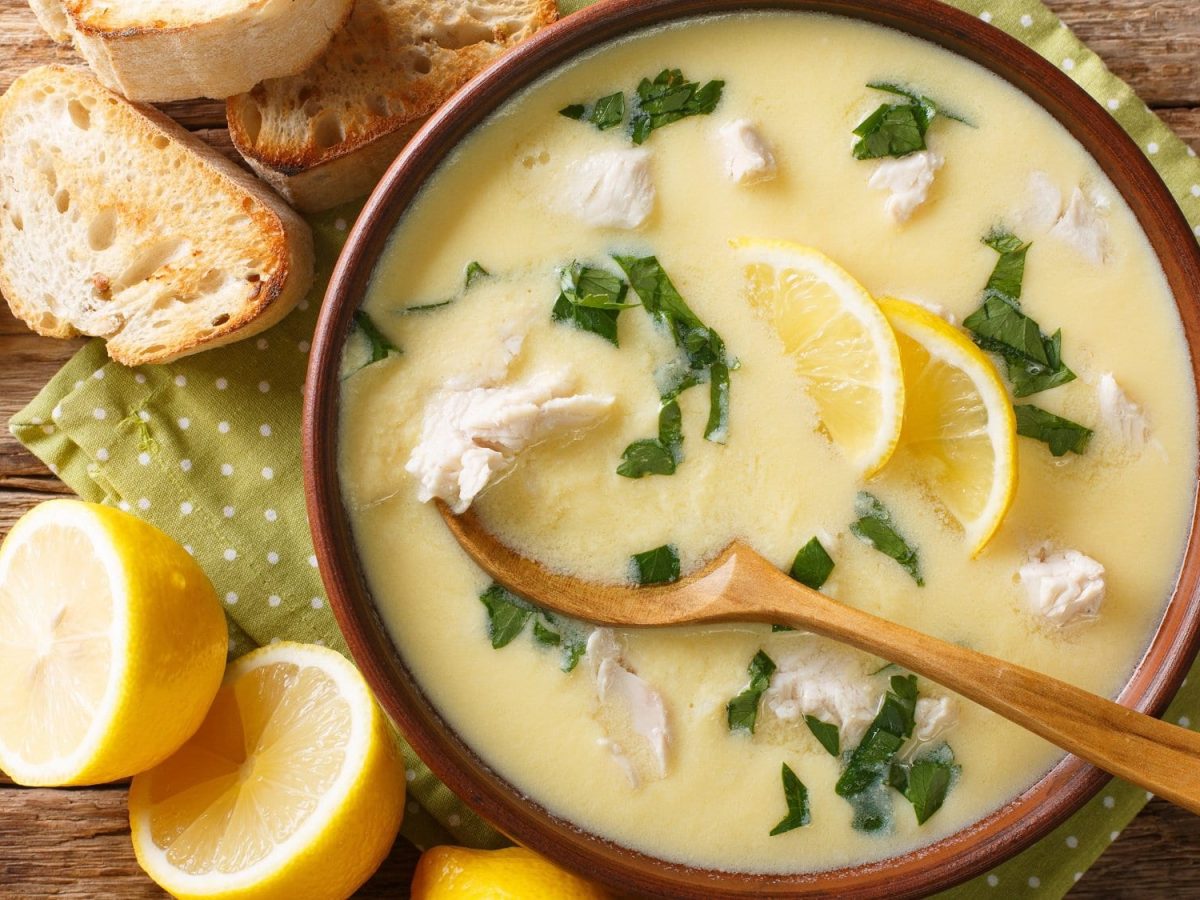 Ein Teller mediterrane Hühnersuppe mit Zitrone und Petersilie auf einem Holztisch. Daneben liegen Zitronenhälften.