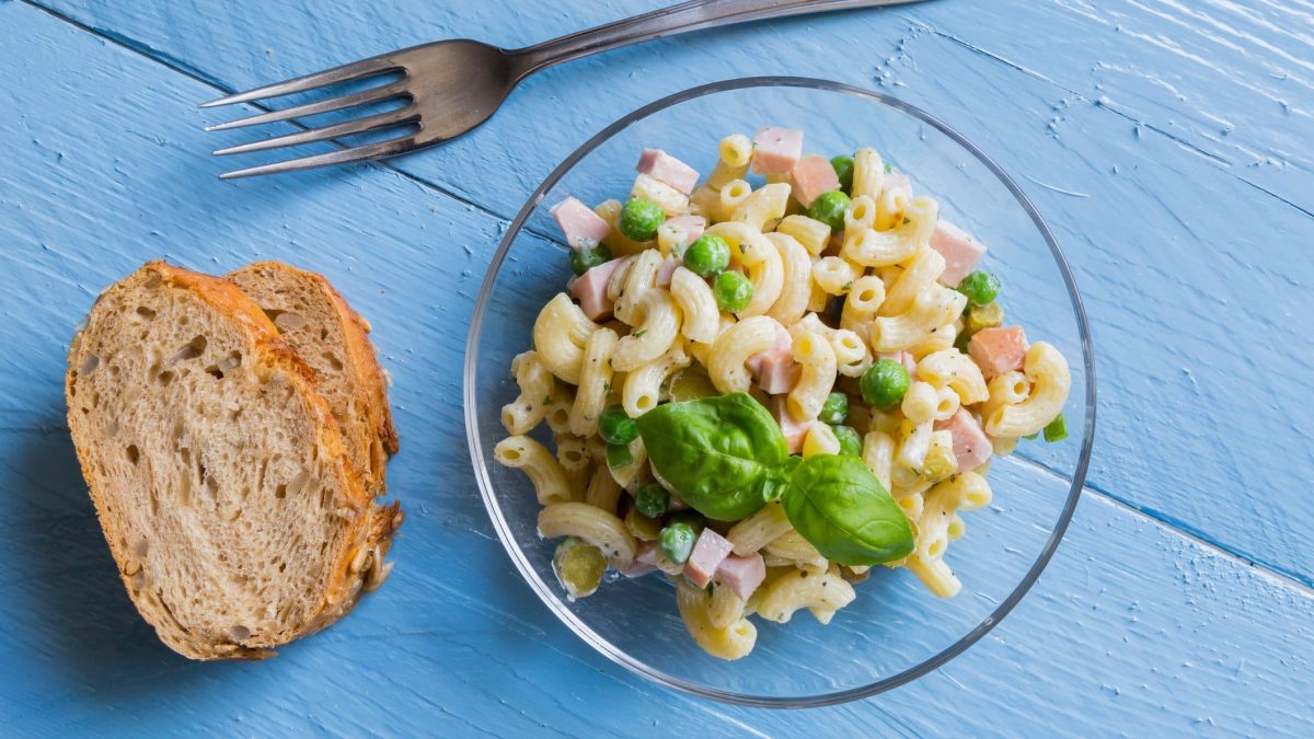 Nudelsalat mit Fleischwurst und Erbsen in einer Glasschale. Diese steht auf einem blauen Holztisch. Daneben Brot und eine Gabel.