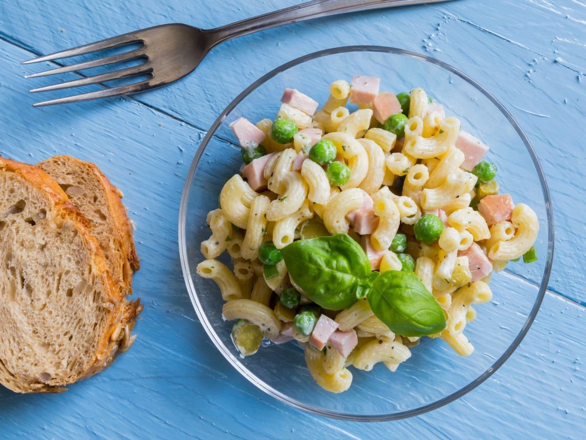 Nudelsalat mit Fleischwurst und Erbsen in einer Glasschale. Diese steht auf einem blauen Holztisch. Daneben Brot und eine Gabel.