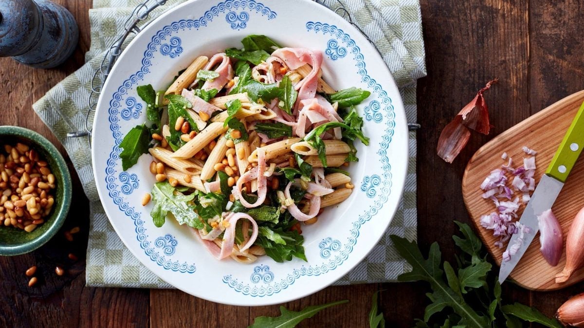 Ein Teller Nudelsalat mit Rucola, Schinken und Pinienkernen in der Draufsicht, daneben die Zutaten.