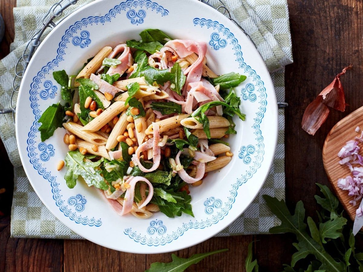 Ein Teller Nudelsalat mit Rucola, Schinken und Pinienkernen in der Draufsicht, daneben die Zutaten.