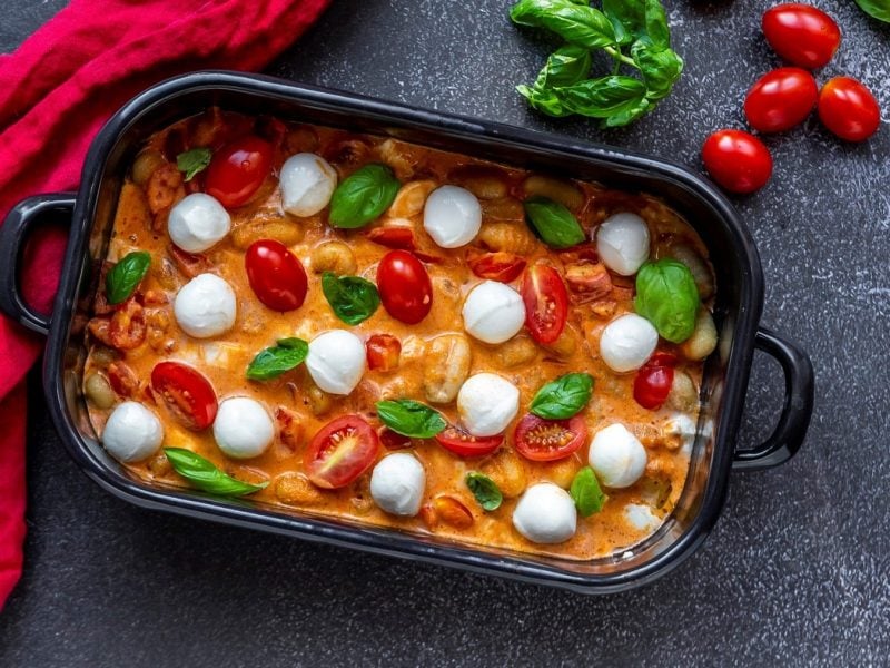 Ofen-Gnocchi mit Tomate & Mozzarella in dunkler Auflaufform. Daneben liegen ein rotes Tuch, Tomaten und einige Blätter Basilikum.