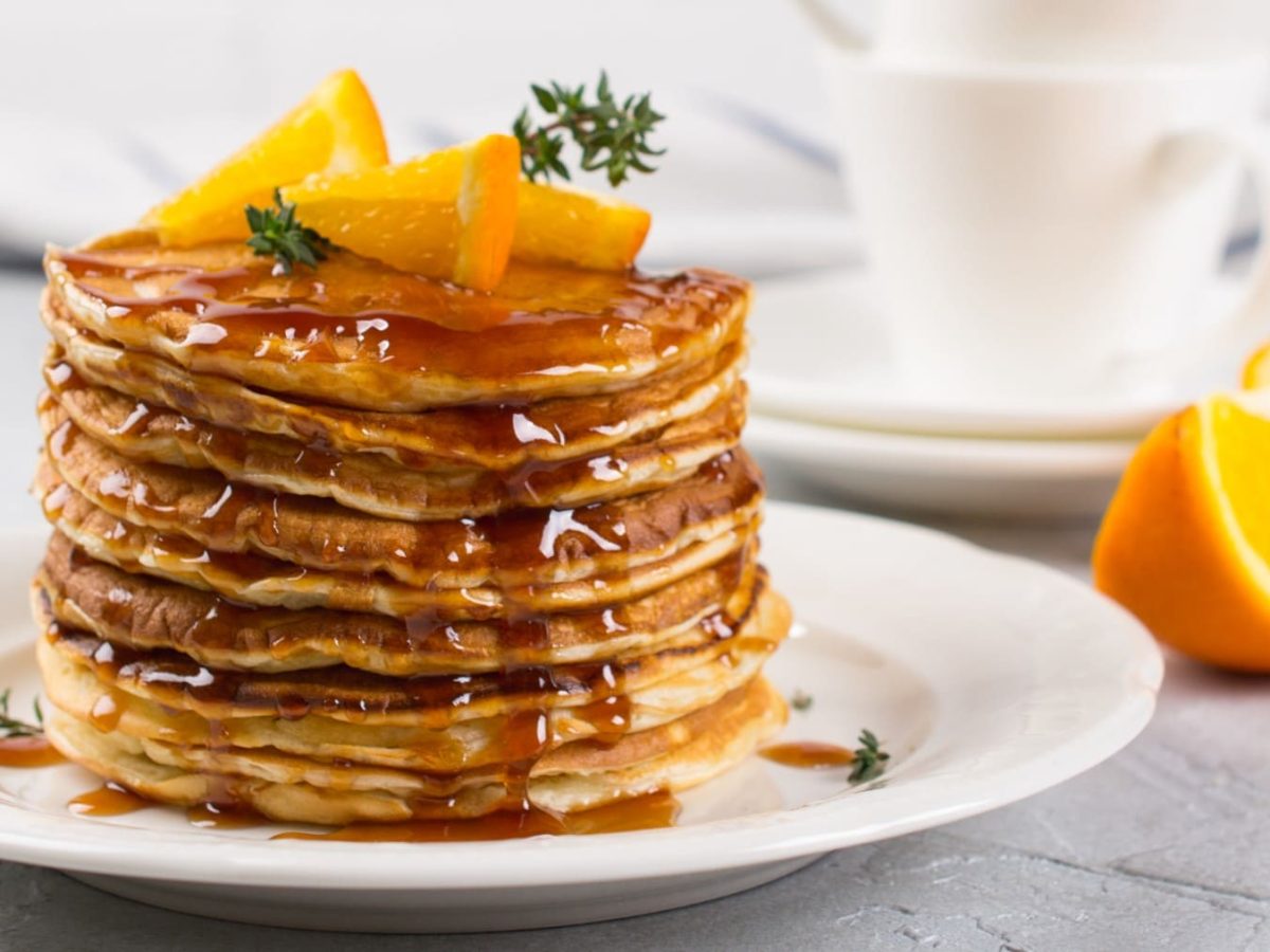 Gestapelte Orangenpancakes mit Ingwersirup und Orangenstückchen auf einem weißen Teller, im hellen Hintergrund Orangenspalten und Geschirr.