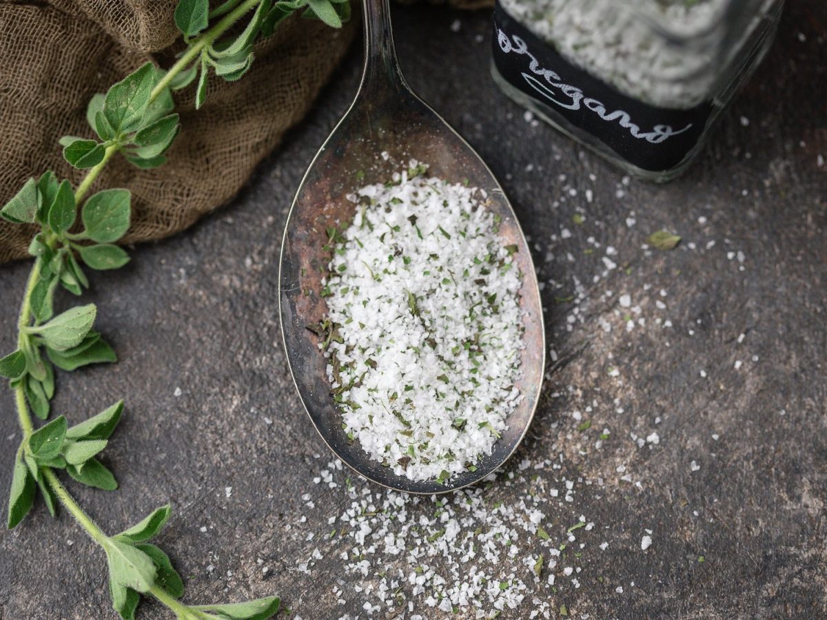 Draufsicht: Ein Löffel selbst gemachtes Oregano-Salz auf einem dunklem Untergrund. Daneben liegt frisches Oregano.