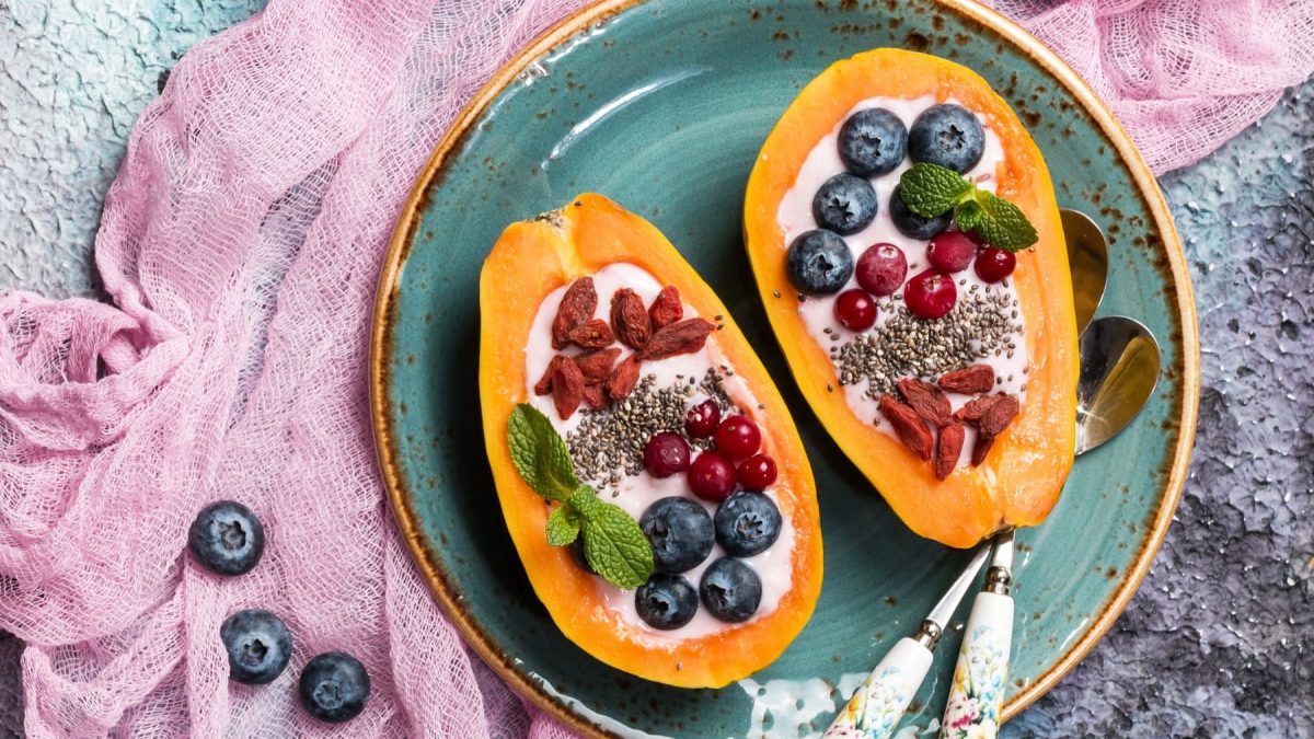 Zwei Papayahälften mit den Toppings auf einem blauen Teller und einem violette-blauen Tisch.