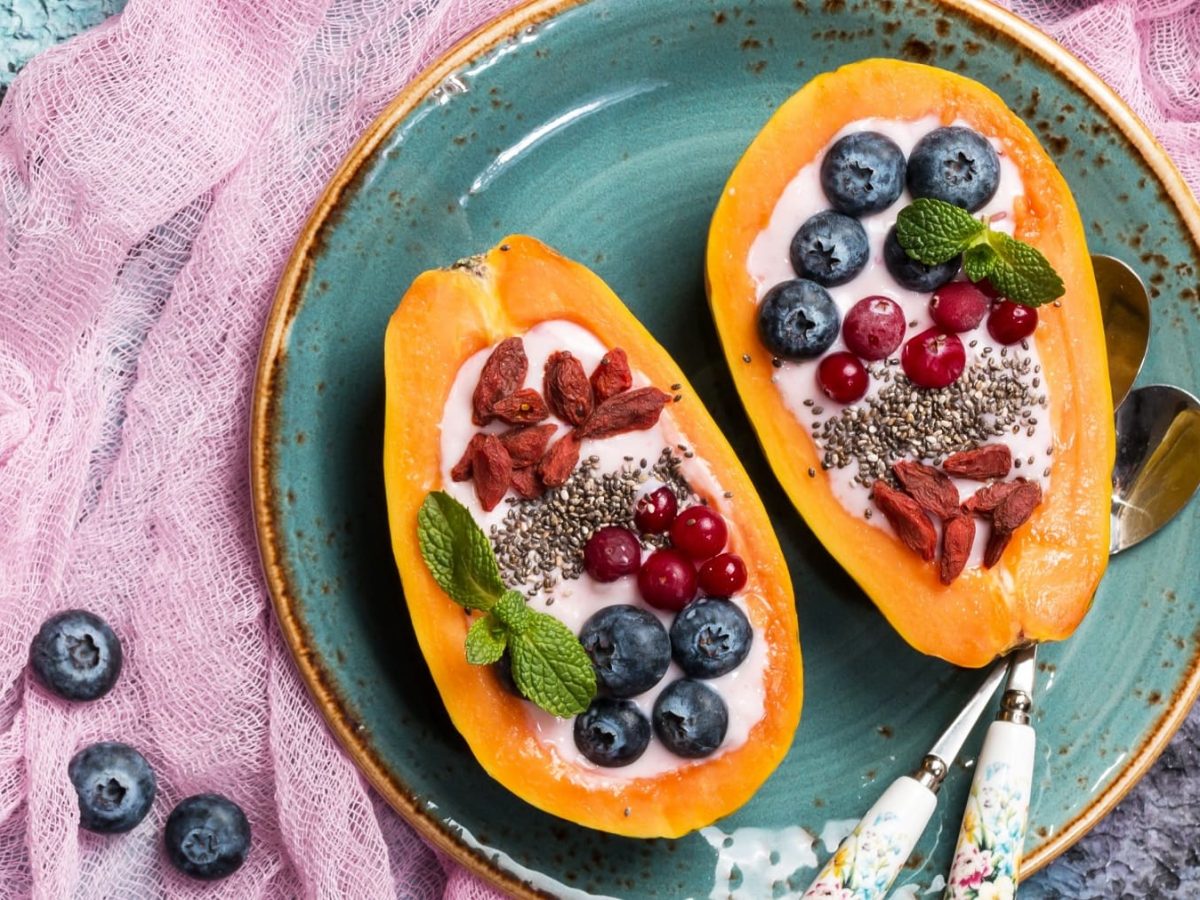 Zwei Papayahälften mit den Toppings auf einem blauen Teller und einem violette-blauen Tisch.