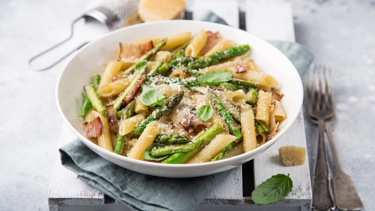 Ein weißer Teller mit Penne, grünem Spargel und Schinken auf einer weißen Holzkiste und grauem Untergrund.