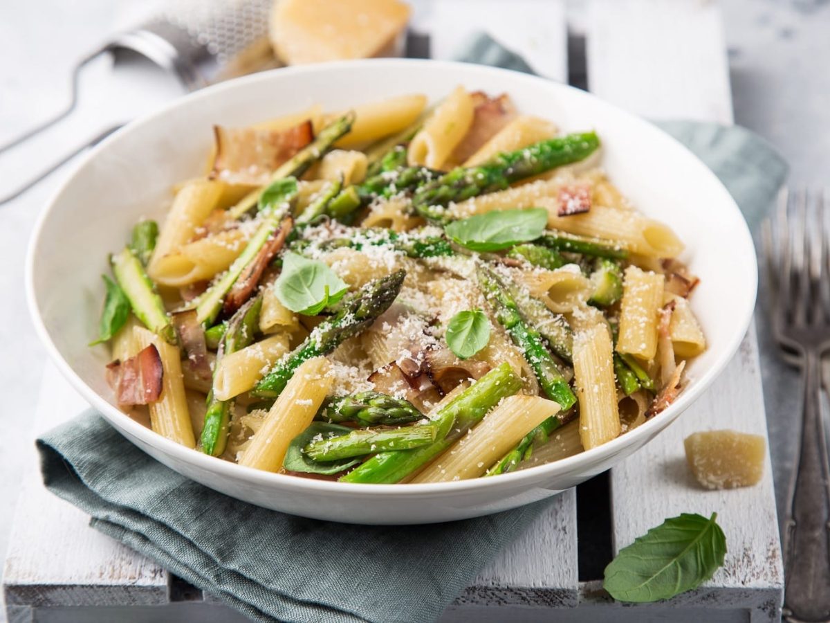 Ein weißer Teller mit Penne, grünem Spargel und Schinken auf einer weißen Holzkiste und grauem Untergrund.