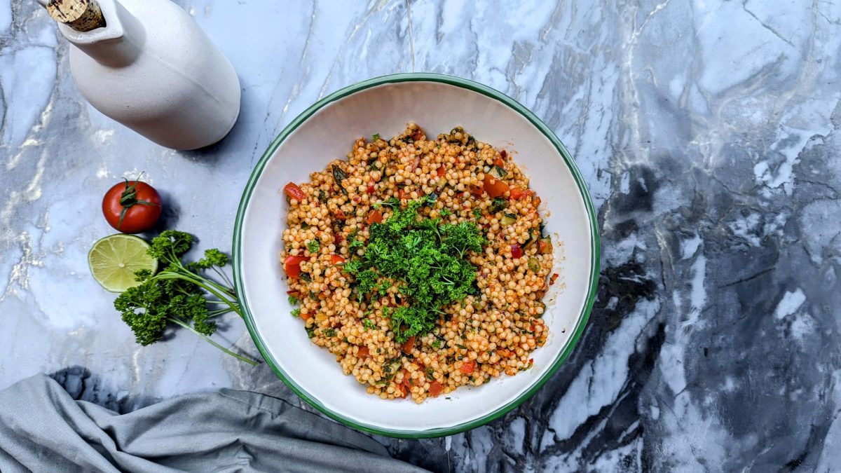 Eine weiße Schüssel mit dem Perlen-Couscous-Salat auf einem Marmortisch mit Limette, Tomate und Petersilie von oben fotografiert.