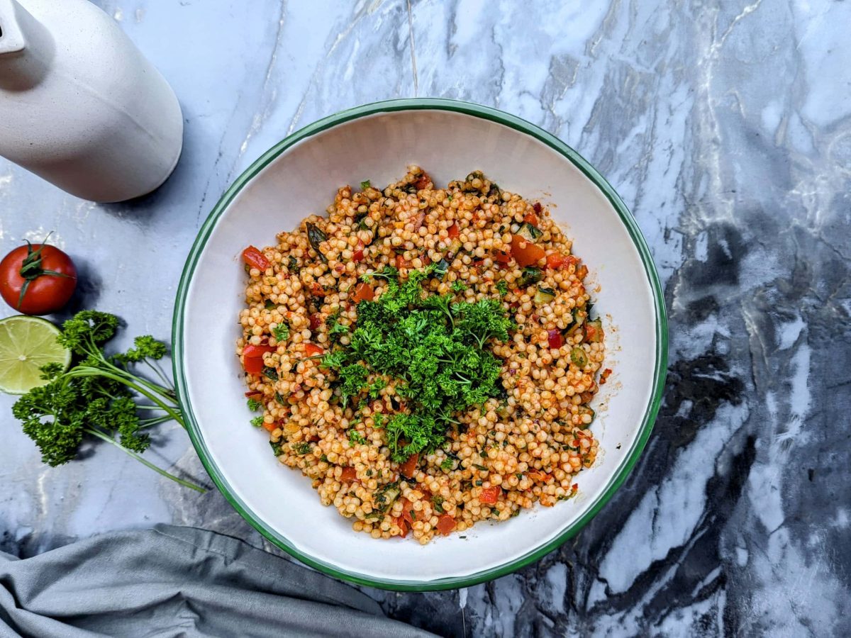 Eine weiße Schüssel mit dem Perlen-Couscous-Salat auf einem Marmortisch mit Limette, Tomate und Petersilie von oben fotografiert.