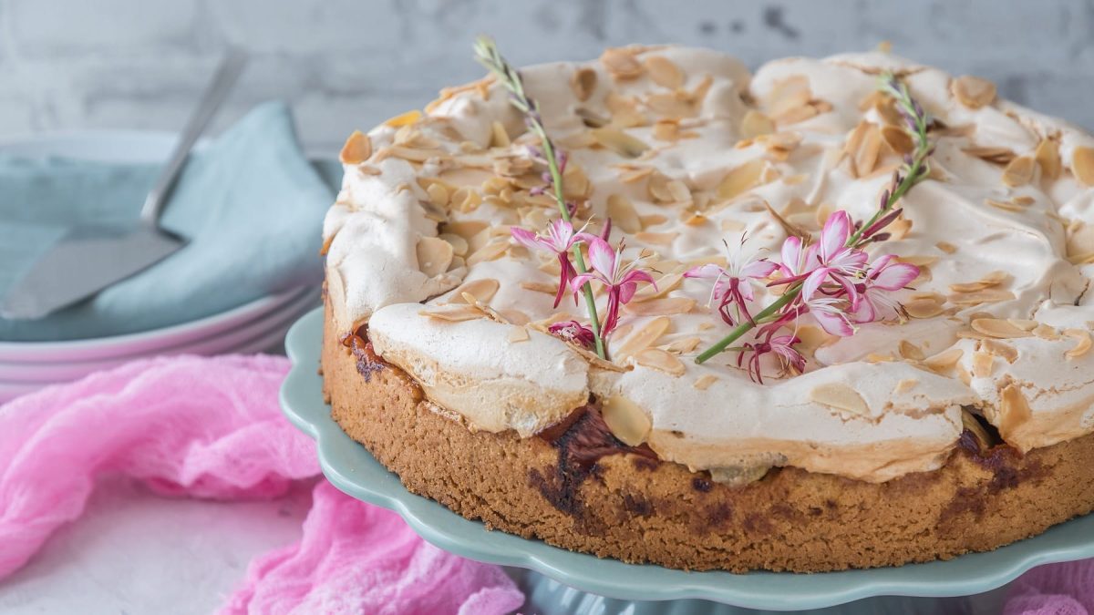 Der Rhabarber-Baiser-Kuchen auf einem hellblauen Tortenständer und grauem Steintisch. Daneben etwas pinker Tüll.