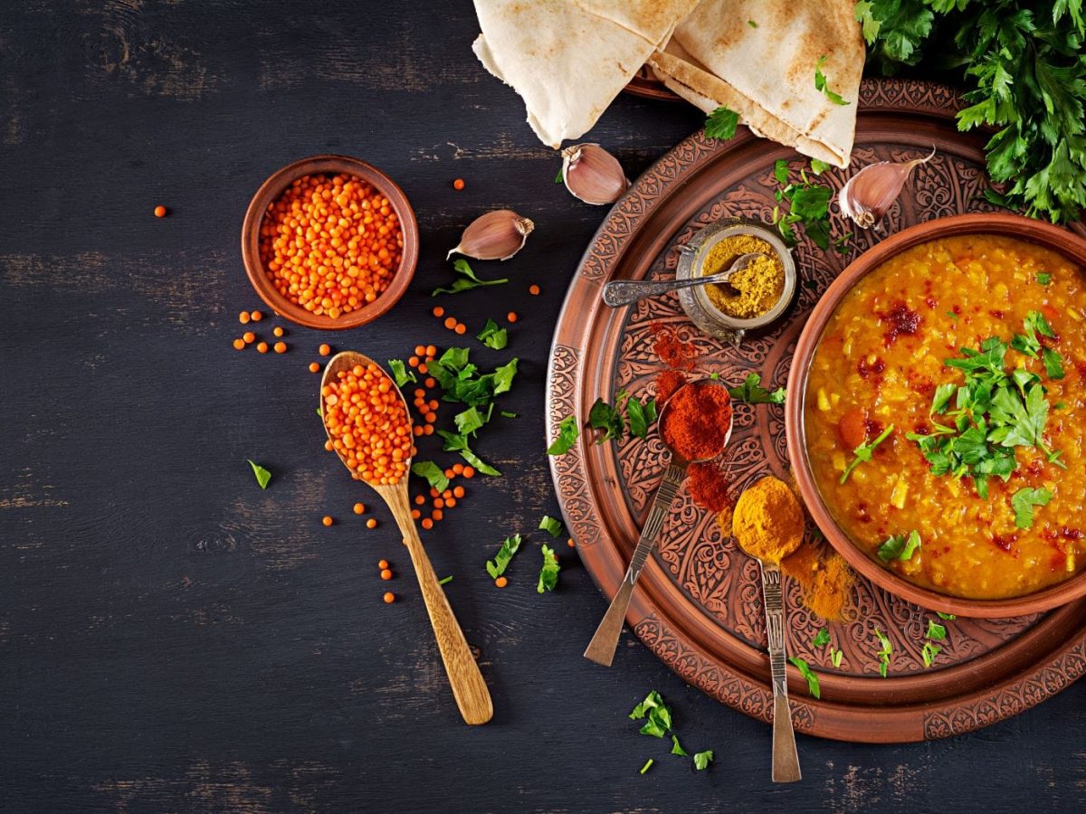 Rhabarber-Curry mit roten Linsen und Kartoffeln in Schale auf braunem Teller auf dunklem Untergrund mit Löffel und Gewürzen. Im Bild ein Löffel mit Masala, eine Schale mit Chilipulver, Koriander und Brot. Draufsicht.