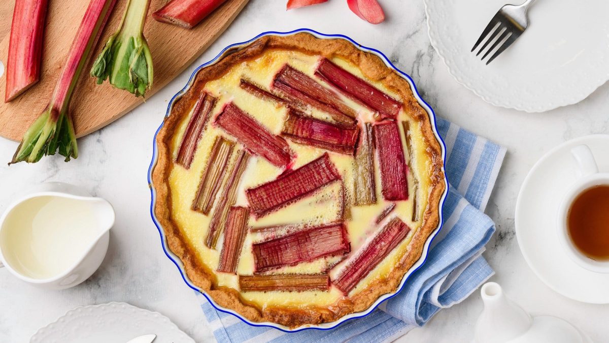 Eine Rhabarber-Pudding-Tarte in der Draufsicht, daneben Rhabarberstangen und Kaffeegedeck.