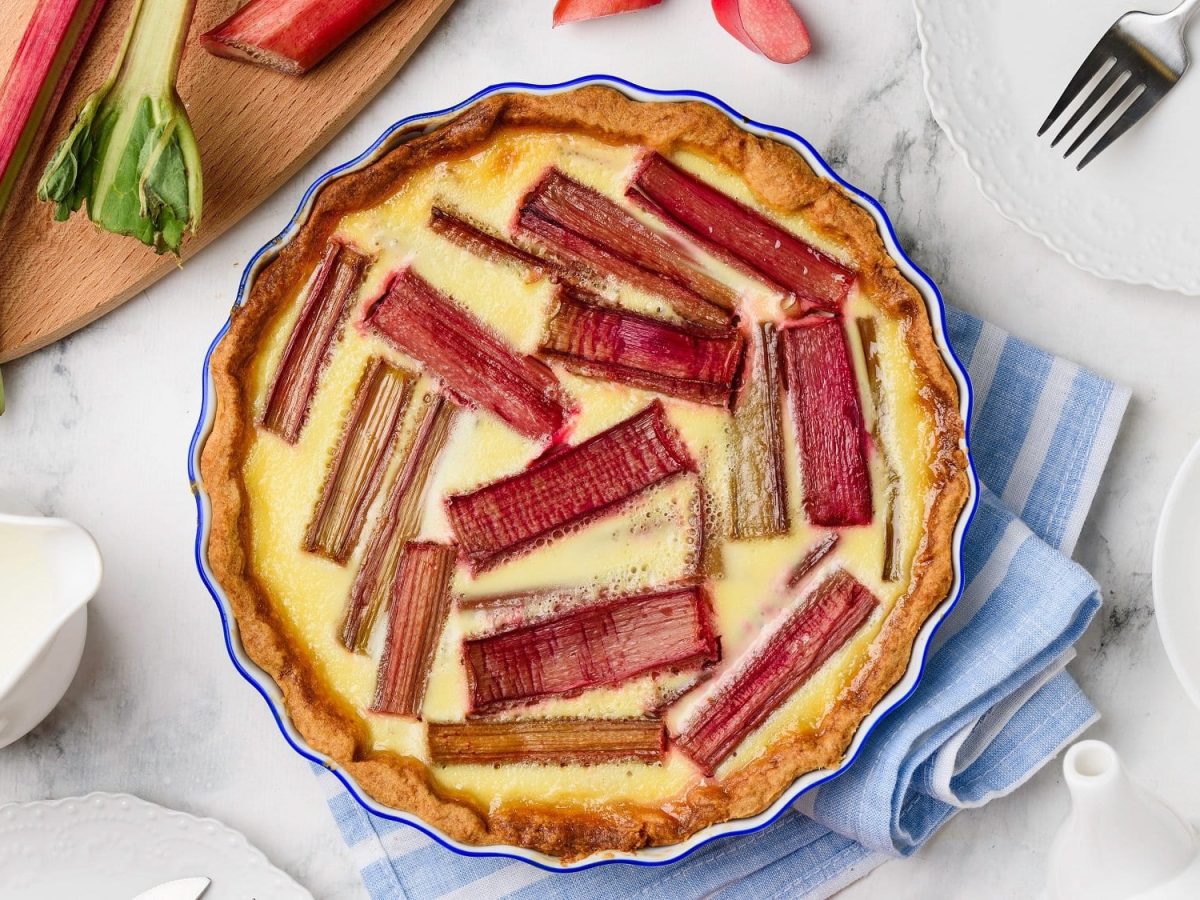 Eine Rhabarber-Pudding-Tarte in der Draufsicht, daneben Rhabarberstangen und Kaffeegedeck.