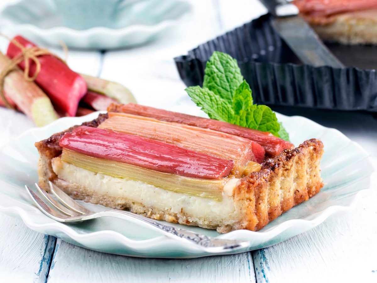 Ein Stück der Rhabarber-Tarte auf einem weißen Teller und einem weißen Holztisch.