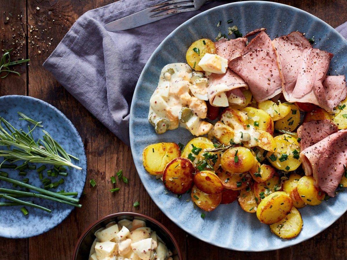 Draufsicht: Ein Teller Kräuter-Kartoffeln mit Roastbeef liegt auf einem dunklen Holztisch. Daneben liegen Rosmarin, kein geschnittenes gekochtes Ei, Pfeffer und ein Tuch.