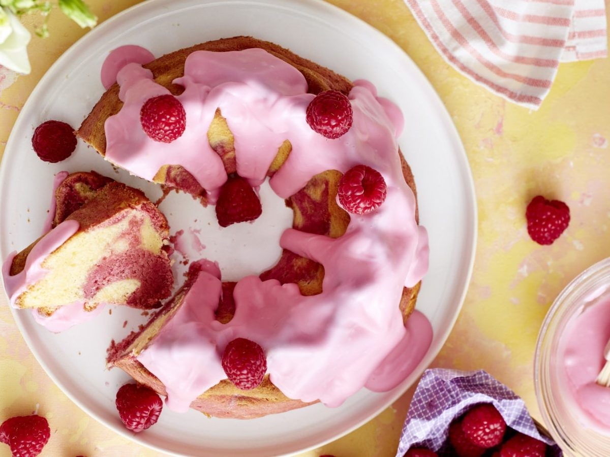 Ein Rosa Marmor-Gugelhupf mit Himbeeren auf einem weißen Teller, auf einem gelben Tischtuch. Rechts unten eine rosa Glasur und vereinzelt ein paar Himbeeren.