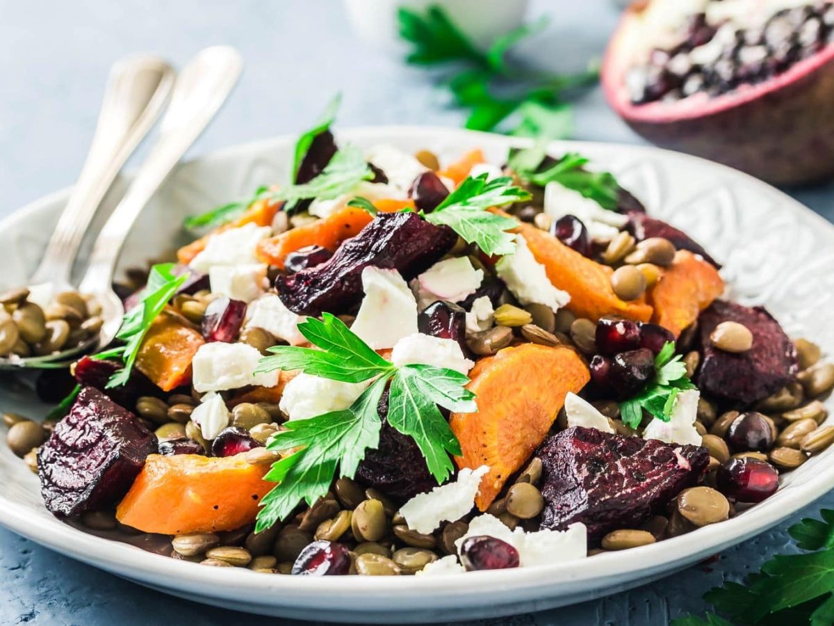 Rote Bete-Linsen-Salat mit Feta, Karotte und Granatapfel mit Besteck auf weißem Teller. Daneben Petersilie und ein halber Granattapfel. Frontalansicht.