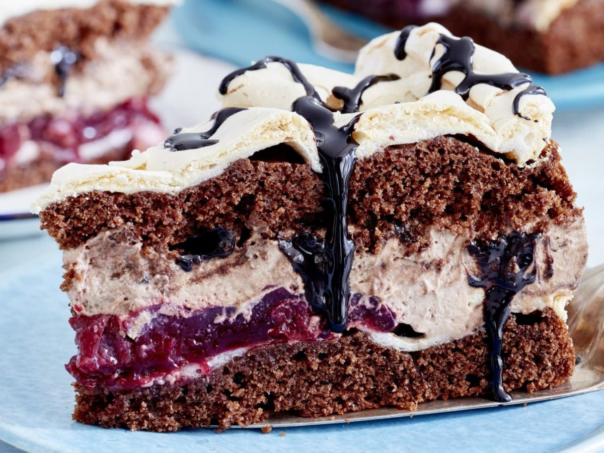 Ein Stück Torte auf einem blauen Teller. Die Torte besteht aus mehreren Schichten. Unten eine Schokoladenkuchenschicht, dann eine Baisermassen, Fruchtschicht, Sahneschicht, Baisermasse, Schokoladenkuchenschicht und noch eine Sahneschicht.