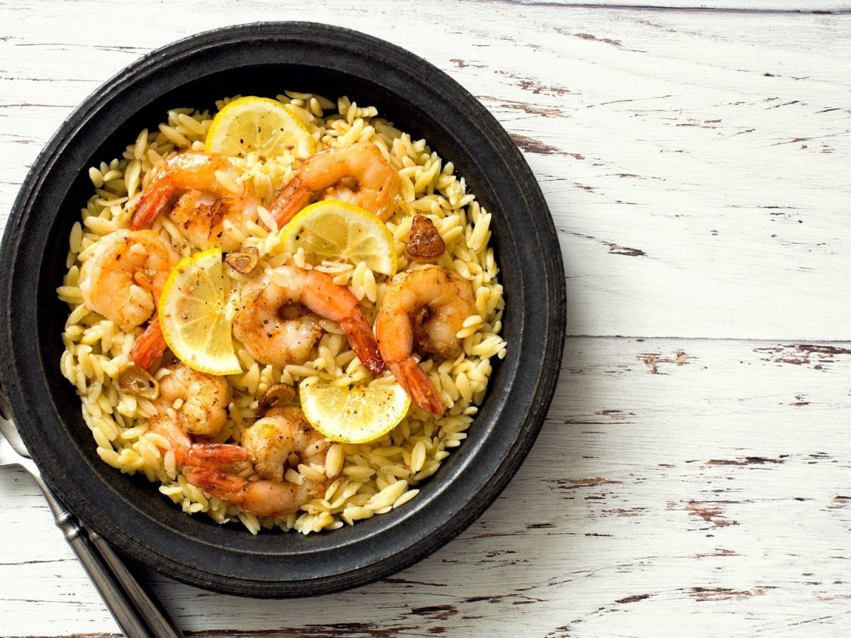 Draufsicht: eine schwarze Schüssel mit Orzo-Garnelen auf einem weißen Untergrund aus Holz: Orzo-Garnelen-Pfanne