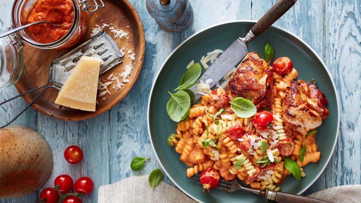 Draufsicht: Schnitzeltaschen mit Pesto-Nudeln und Basilikum auf einem grünen Teller, daneben Parmesan, rotes Pesto und einzelne Tomaten auf blauem Holzuntergrund.