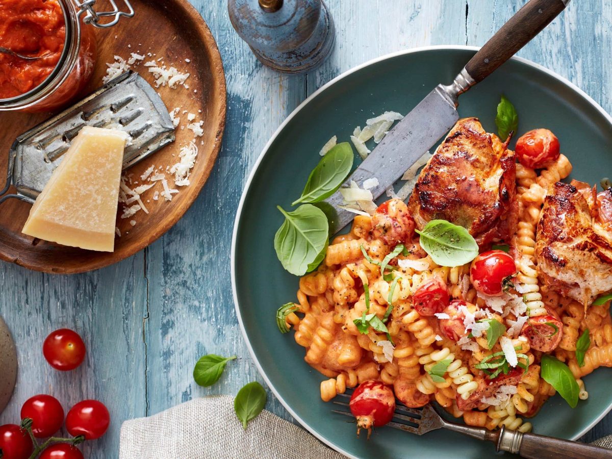 Draufsicht: Schnitzeltaschen mit Pesto-Nudeln und Basilikum auf einem grünen Teller, daneben Parmesan, rotes Pesto und einzelne Tomaten auf blauem Holzuntergrund.