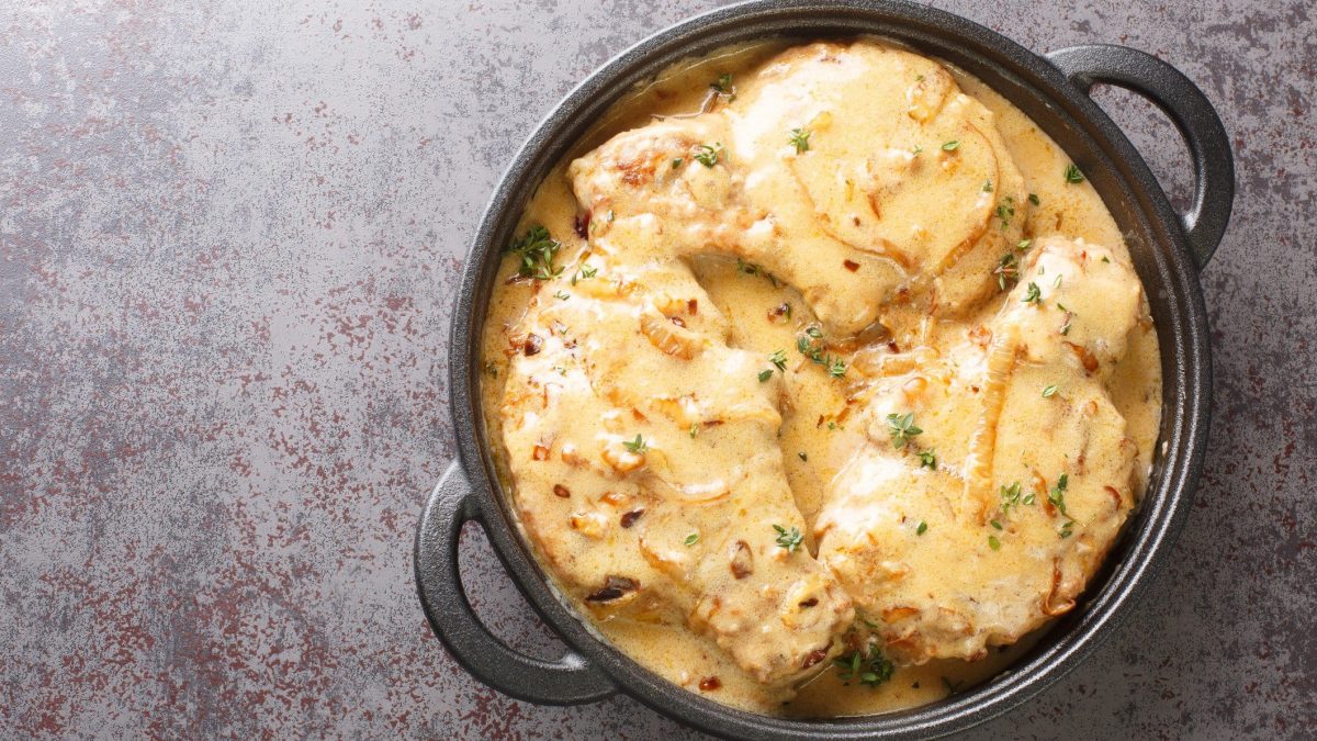 Smothered Pork Chops mit Zwiebel-Sahne-Sauce in schwarzer ofenfester Pfanne auf grauem Untergrund. Draufsicht.