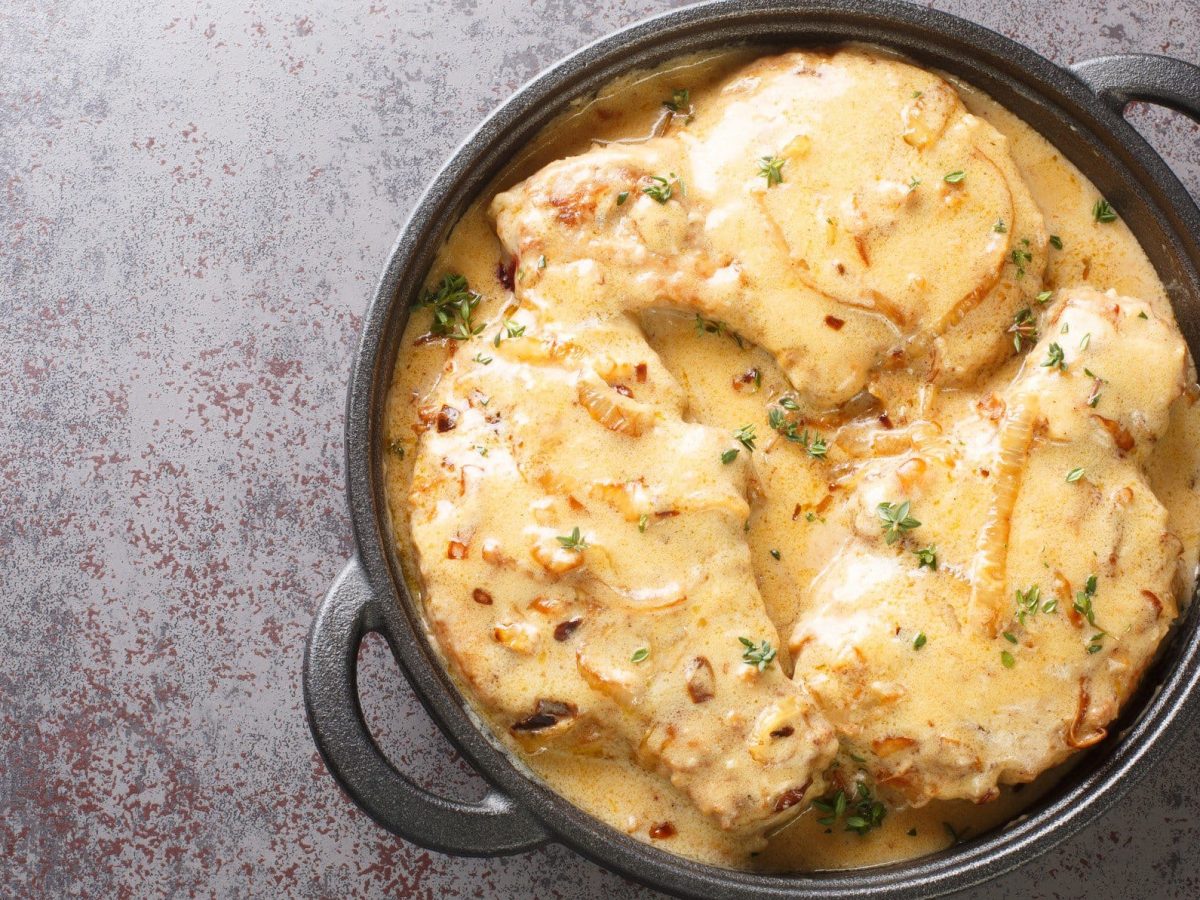 Smothered Pork Chops mit Zwiebel-Sahne-Sauce in schwarzer ofenfester Pfanne auf grauem Untergrund. Draufsicht.