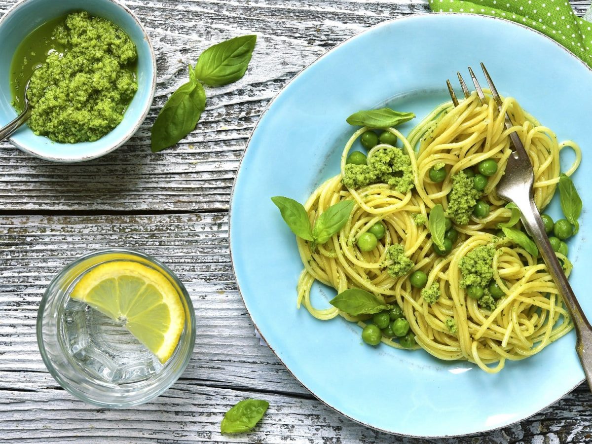 Draufsicht: ein hellblauer Teller mit Spaghettini mit Erbsen und Pesto auf einem Holztisch. Daneben stehen ein Glas Zitronenwasser und eine Schale mit Pesto.