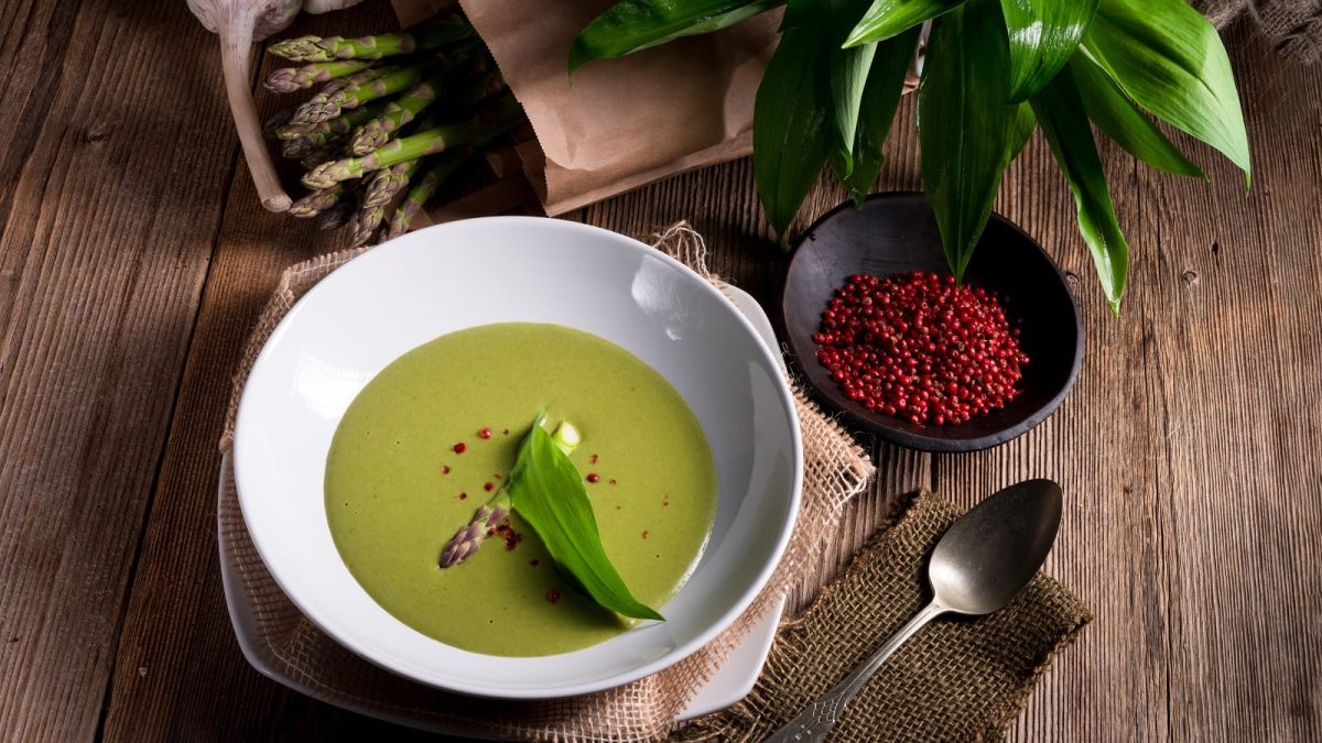 Ein weißer Suppenteller mit Spargelsuppe mit Bärlauch neben frischen Zutaten auf einem Holztisch.