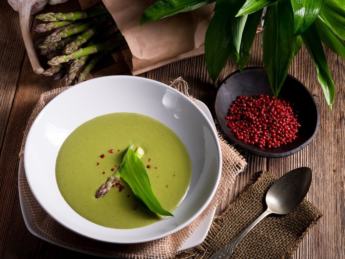 Ein weißer Suppenteller mit Spargelsuppe mit Bärlauch neben frischen Zutaten auf einem Holztisch.