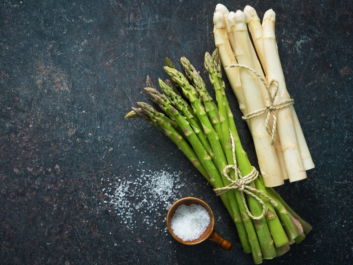 Je ein Bund weißer und grüner Spargel auf einem dunklen Untergrund mit einem kleinen Salzfässchen von oben fotografiert.