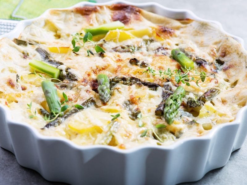 Spargel-Kartoffel-Gratin mit Parmesankruste und mediterranen Kräutern in weißer Kasserole auf grauem Untergrund mit Kräutern. Draufsicht.