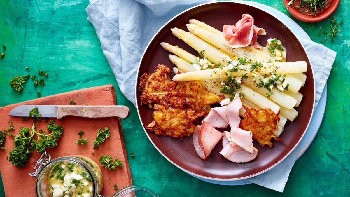 Spargel mit Schinken und Rösti angerichtet auf einem dunkelroten Teller auf einem hellblauen Tuch und einem türkisem Hintergrund. Daneben ein Schneidebrett mit Kräuterbutter und einem Messer.