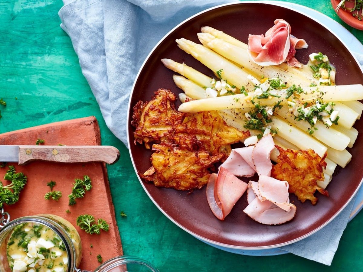 Spargel mit Schinken und Rösti angerichtet auf einem dunkelroten Teller auf einem hellblauen Tuch und einem türkisem Hintergrund. Daneben ein Schneidebrett mit Kräuterbutter und einem Messer.