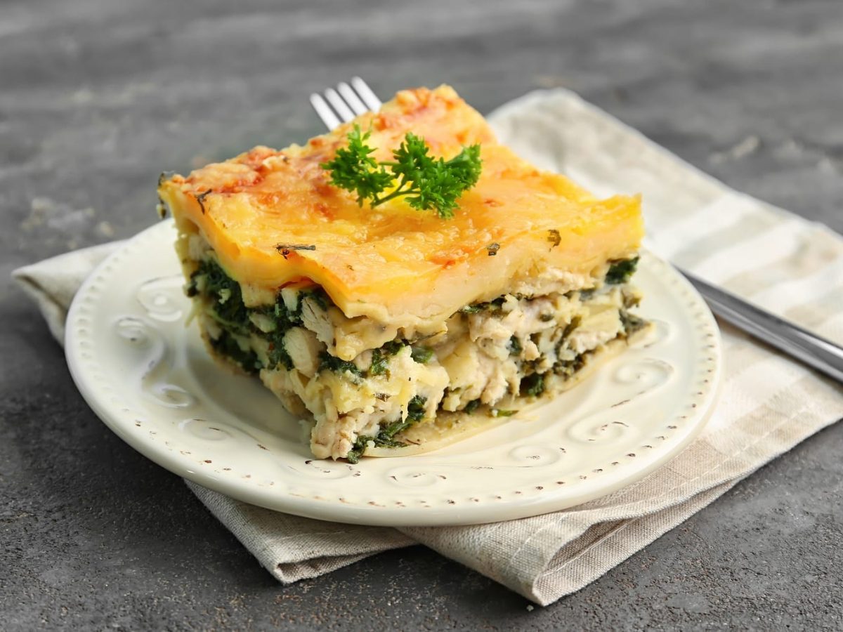 Ein weißer Teller mit einem Stück der Spinat-Lasagne auf grauem Untergrund mit einer hellen Serviette und silbernen Gabel.