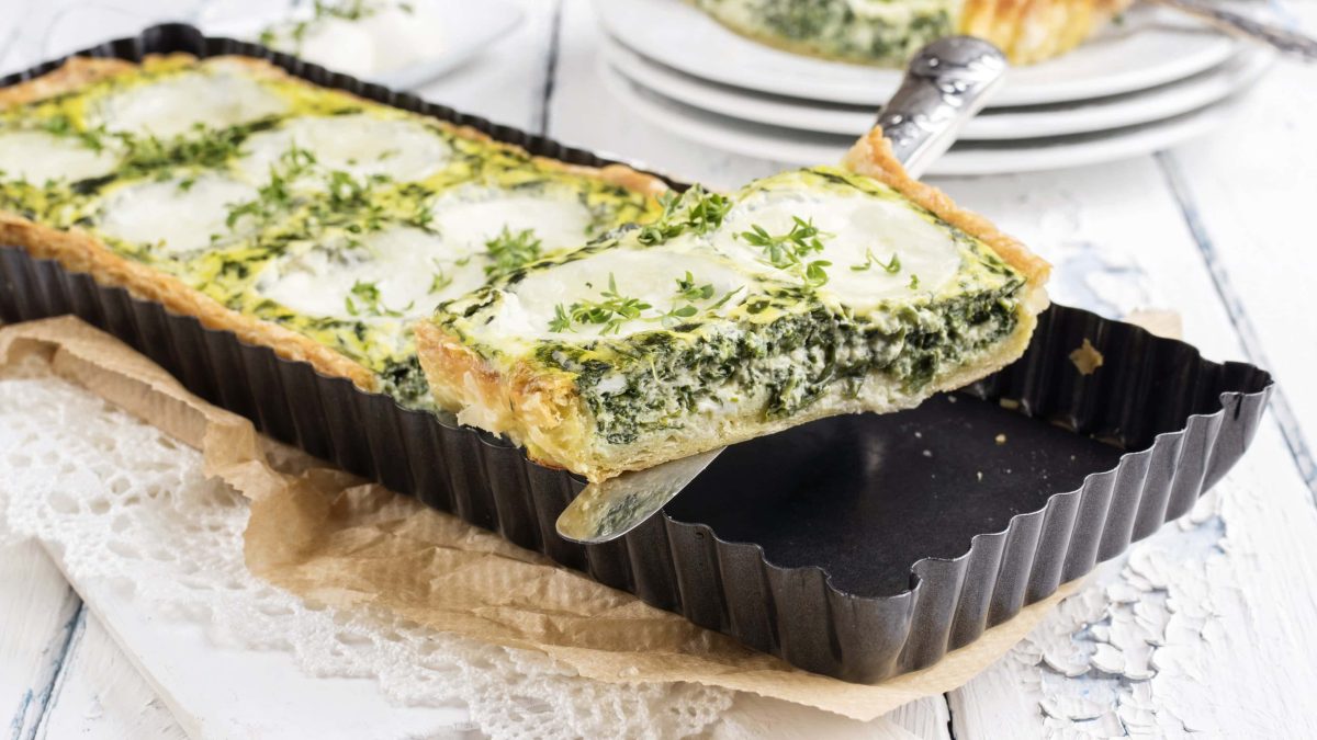 Eine schwarze Auflaufform mit der Spinat-Tarte und Mozzarella auf etwas Backpapier und einem weißen Holztisch.