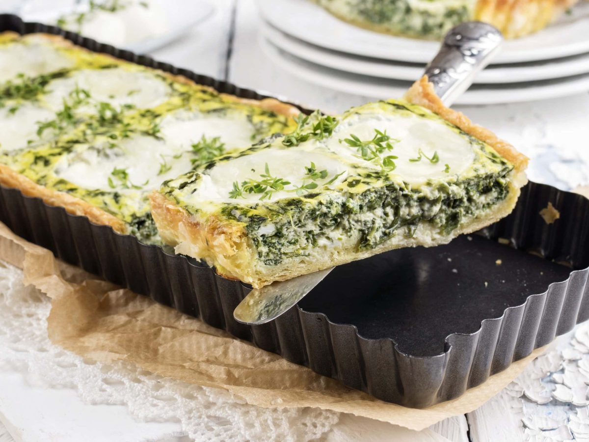 Eine schwarze Auflaufform mit der Spinat-Tarte und Mozzarella auf etwas Backpapier und einem weißen Holztisch.