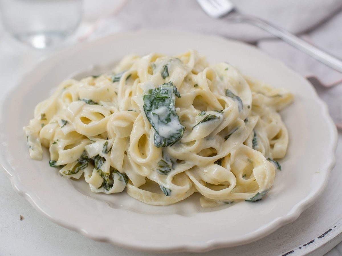 Ein weißer Pastateller mit Tagliatelle mit Bärlauchsahnesauce auf einem weißen Tisch.