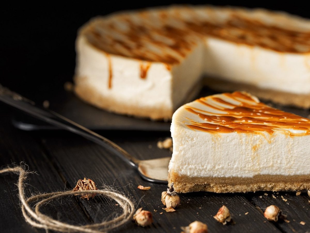 Ein Stück Tahini-Cheesecake mit Salzkaramellsauce, dahinter sieht man unscharf den angeschnittenen Kuchen.