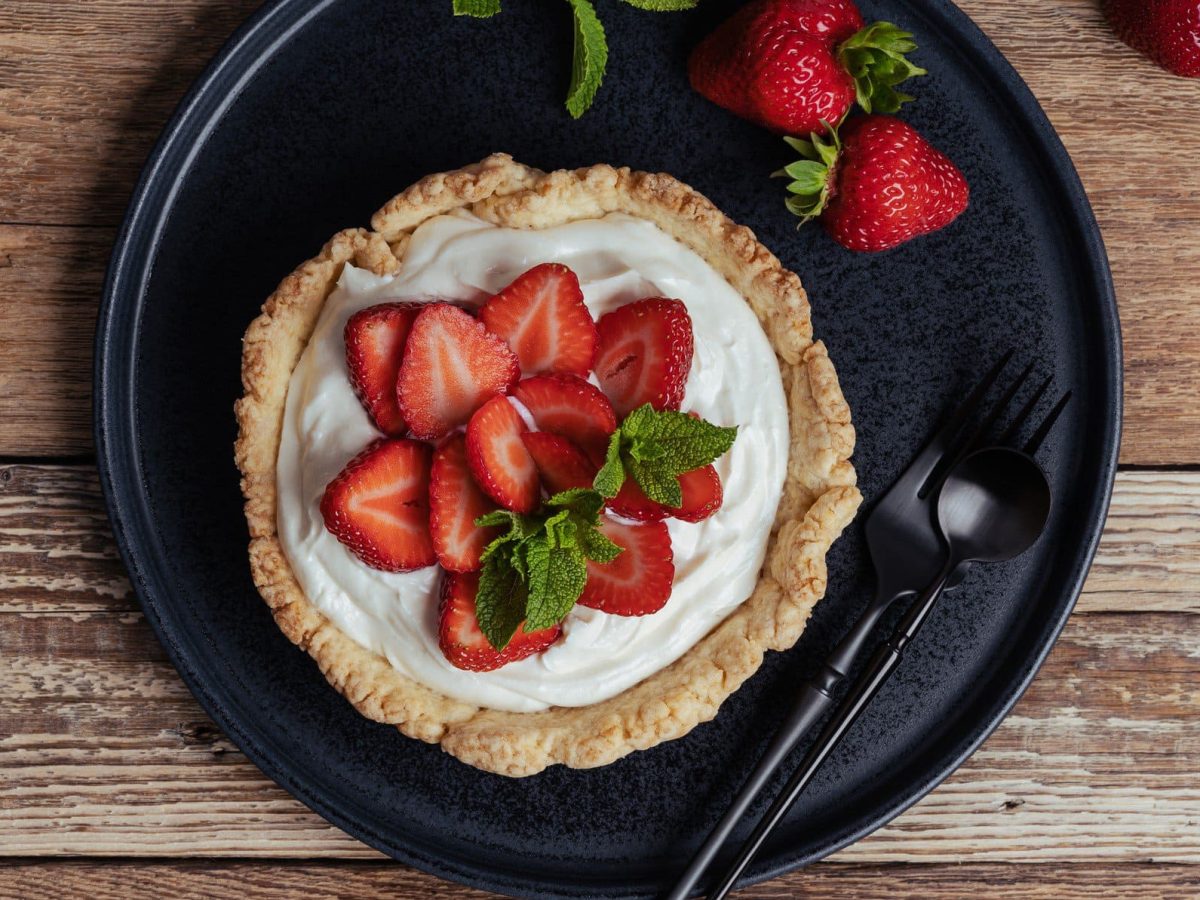 Gefüllte Tarte mit Vanillecreme und marinierten Erdbeeren mit Minzblättern und Erdbeeren auf schwarzem Teller auf Holz. Daneben schwarzes Besteck. Draufsicht.