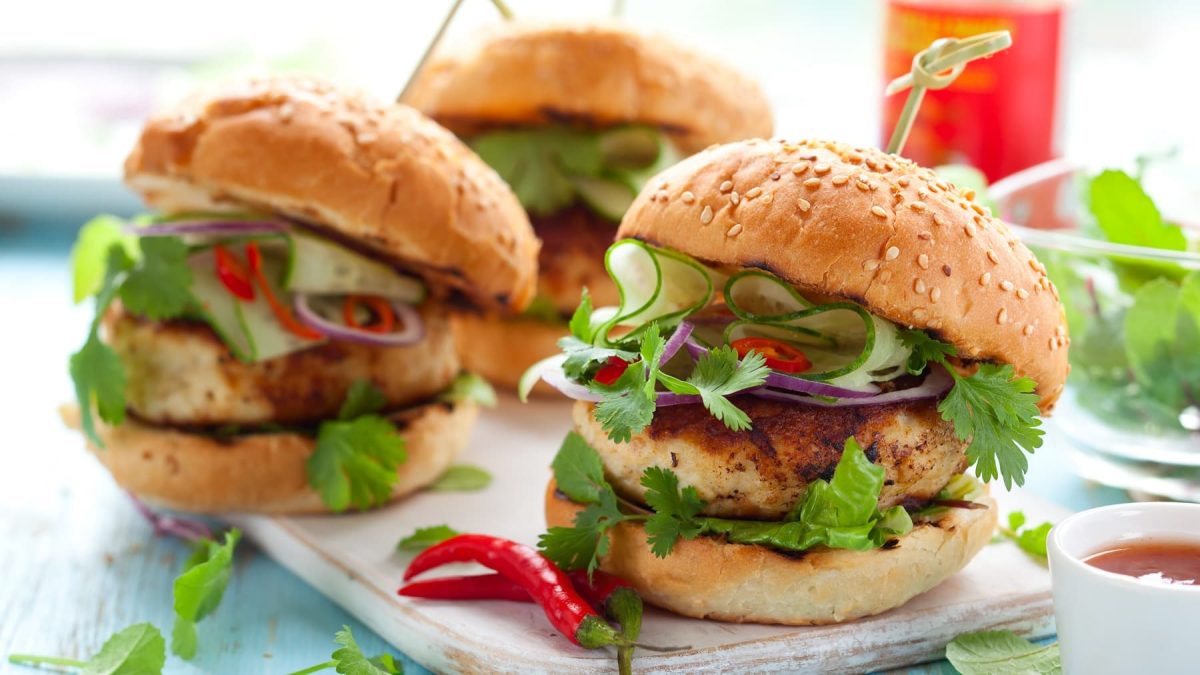 Drei Thai-Chicken-Burger auf einem hellblauen Holztisch.