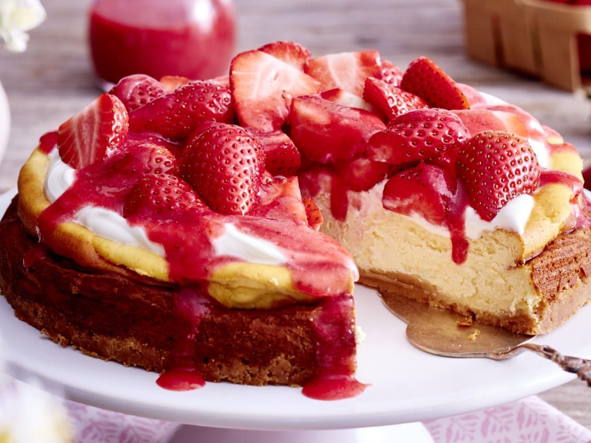 Ein Cheesecake mit Erdbeeren und ein Tortenheber auf einem weißen Teller. Links oben eine kleine Vase mit Blumen, rechts oben eine Packung Erdbeeren.