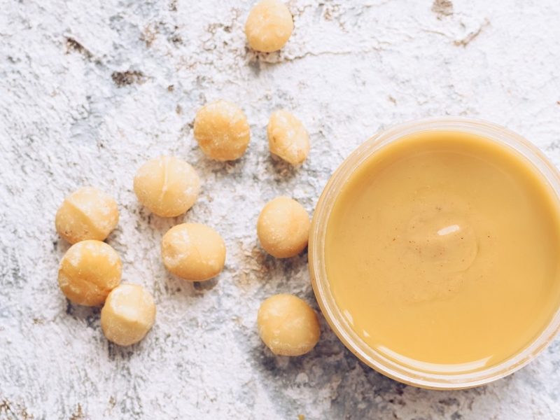 Ein Glas mit Vanille-Macadamia-Butter neben Macadamia-Nüssen.