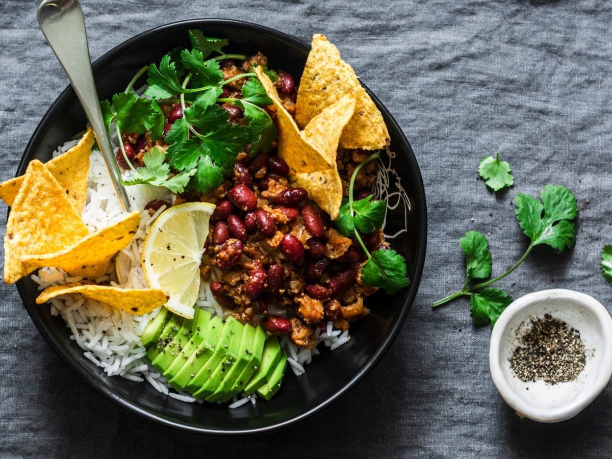 Draufsicht: Vegane Burrito-Bowl mit Tortilla-Chips und einer Gabel in einer schwarzen Schüssel auf grauem Stoffuntergrund, daneben eine Schale schwarzer Pfeffer und etwas Koriander.