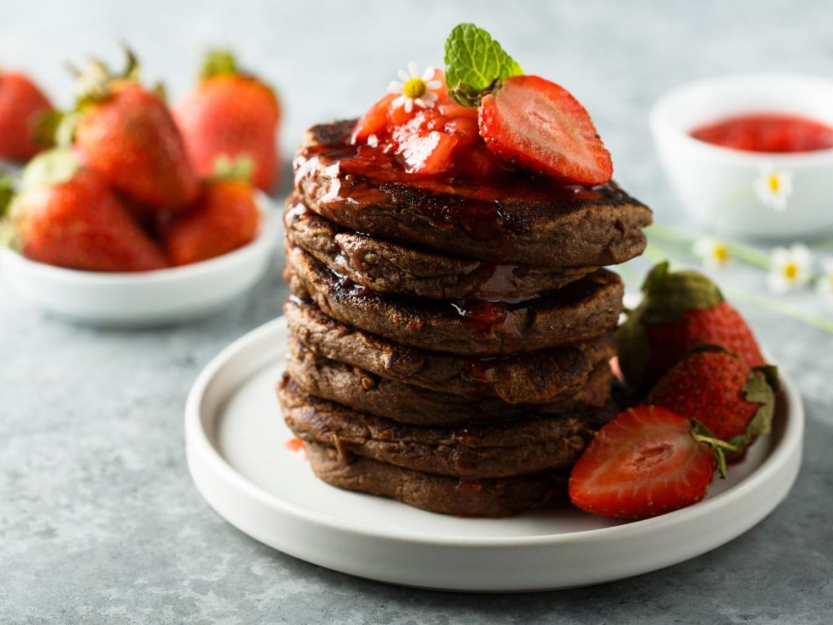 Vegane Schoko-Pancakes mit Erdbeersauce und frischen Erdbeeren auf einem weißen Teller, im Hintergrund mehr Erdbeeren, alles auf hellgrauem Untergrund.