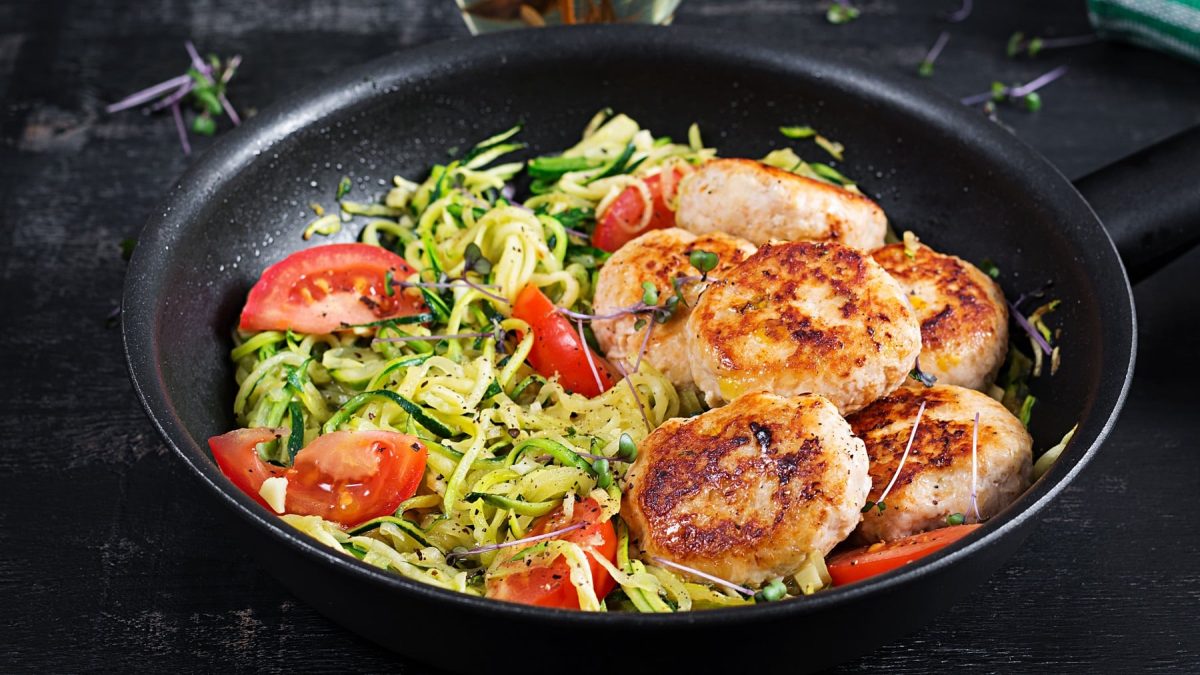 Eine schwarze Pfanne mit den Zoodles und Hähnchenfrikadellen auf einem schwarzen Untergrund.