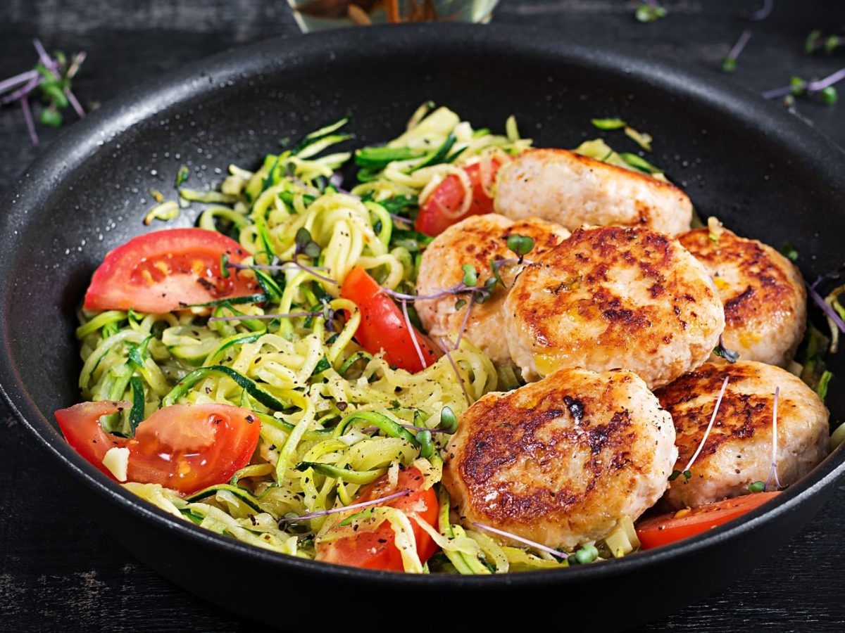 Eine schwarze Pfanne mit den Zoodles und Hähnchenfrikadellen auf einem schwarzen Untergrund.